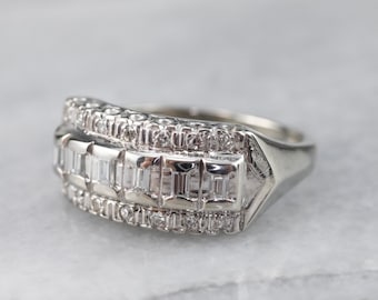 May include: A white gold ring with a rectangular design. The ring features a row of rectangular-cut stones in the center, flanked by rows of smaller, round stones. The ring is set on a white surface.