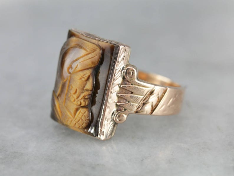 May include: A gold ring with a carved tiger's eye stone featuring a detailed profile of a person. The band is engraved with a floral design.