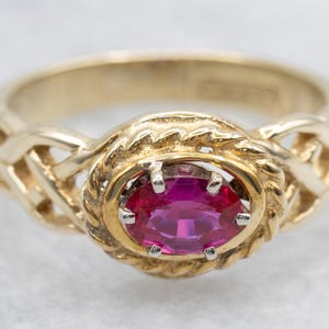May include: A gold ring featuring a vibrant oval-cut ruby gemstone. The ruby is set in a gold bezel, surrounded by a textured, rope-like design. The band has an intricate, woven pattern.