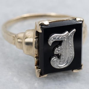 May include: A gold-toned ring with a rectangular black onyx stone. The stone features a silver-colored letter "J" in an ornate, stylized font. The ring has a classic, vintage design.