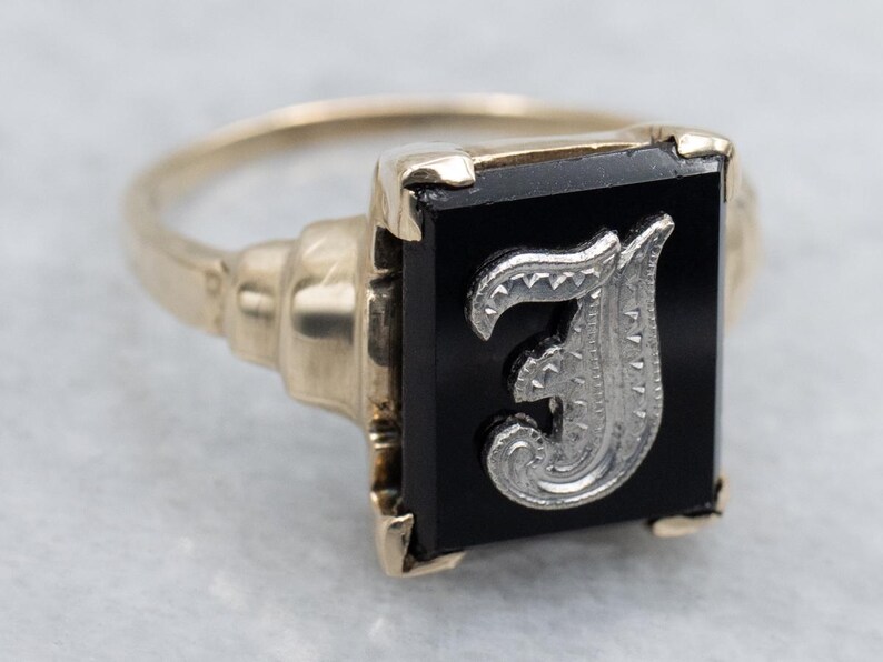 May include: A gold-toned ring with a rectangular black onyx stone. The stone features a silver-colored letter "J" in an ornate, stylized font. The ring has a classic, vintage design.