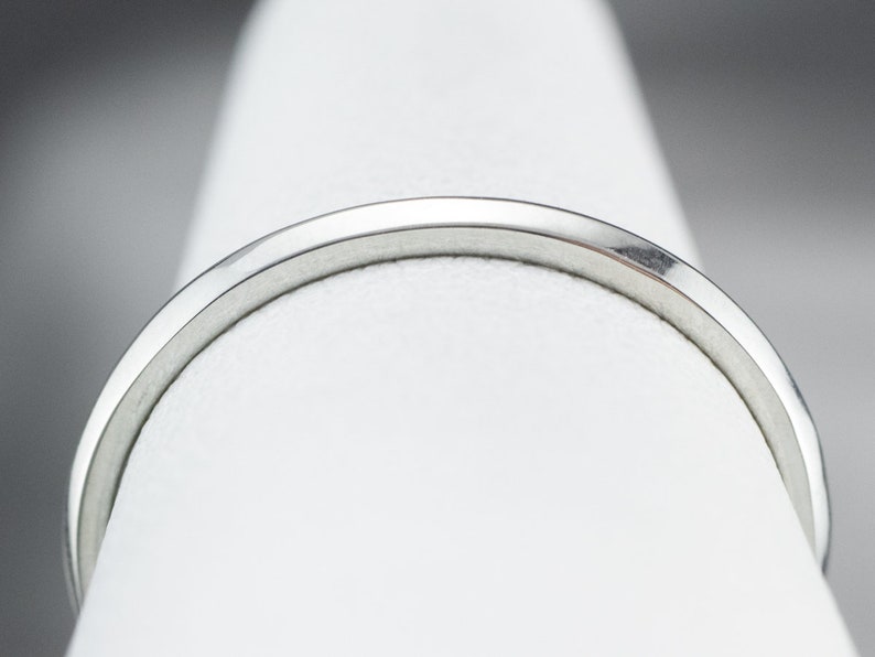 May include: A close-up of a silver ring with a smooth, polished finish. The ring is on a white display stand.