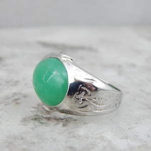 May include: A silver ring with a green gemstone set in the center. The band is decorated with an intricate floral design.