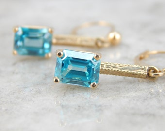 May include: A pair of gold dangle earrings with blue gemstone drops. The earrings have a delicate, intricate design.