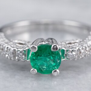 May include: A silver ring featuring a large emerald gemstone surrounded by smaller diamonds. The band is set with a row of diamonds.