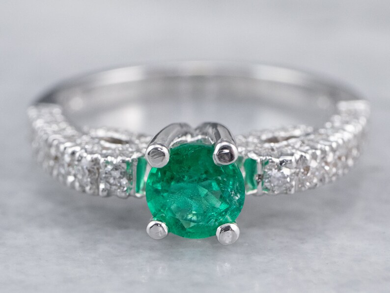 May include: A silver ring featuring a large emerald gemstone surrounded by smaller diamonds. The band is set with a row of diamonds.