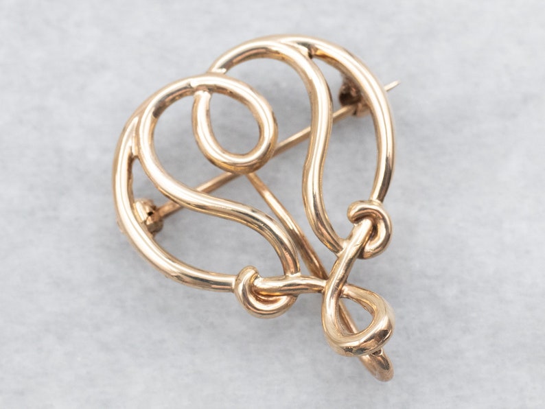 May include: A gold-toned, abstract heart-shaped brooch with a delicate, intricate design. The brooch is made of a single piece of wire that is twisted and turned to create the shape.