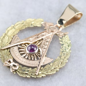 May include: Gold pendant featuring a compass and square design, encircled by a wreath of leaves. A pink gemstone is set in the centre of the design.