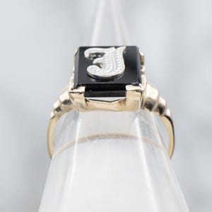 May include: A gold ring with a rectangular black onyx stone. The stone features a silver letter "J". The ring has a classic design with a detailed band. A piece of jewelry.