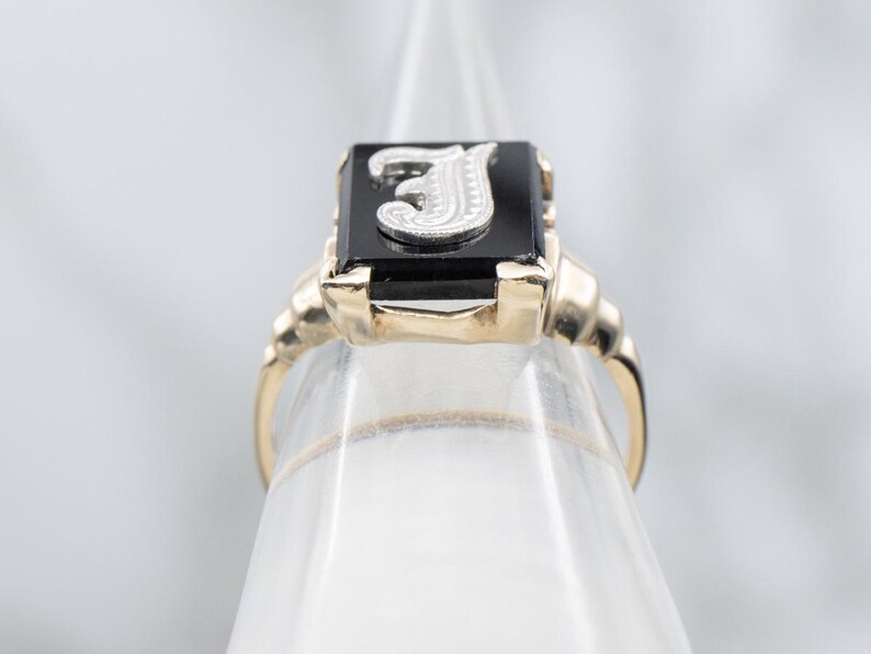May include: A gold ring with a rectangular black onyx stone. The stone features a silver letter "J". The ring has a classic design with a detailed band. A piece of jewelry.