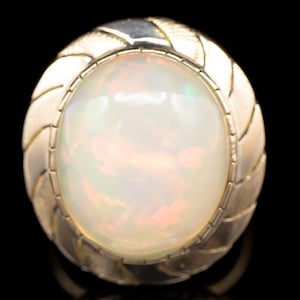 Peut inclure: Une bague dorée ornée d'une grande opale ovale. L'opale présente une base blanc laiteux avec de subtils reflets de couleurs. Le bandeau de la bague a un motif tissé texturé autour de la pierre, créant une esthétique vintage.