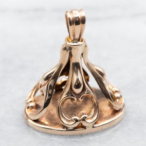 May include: Gold-toned metal seal with an ornate design. The seal has a loop at the top for attaching to a chain.