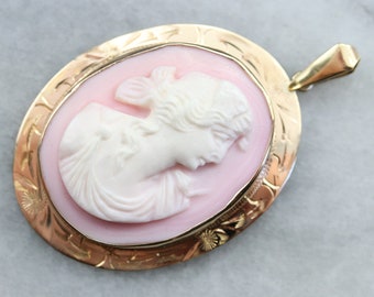 Pode incluir: Um pingente dourado com um camafeu de perfil de mulher. O camafeu está engastado em um oval rosa e rodeado por uma moldura dourada detalhada.