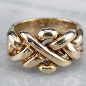 Puede incluir: Un anillo de puzzle dorado con un diseño complejo entrelazado. El anillo es grueso y ancho, con un acabado liso y pulido.