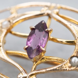 May include: A gold brooch with a purple gemstone and pearl accents. The brooch is in an intricate design with a delicate filigree pattern.