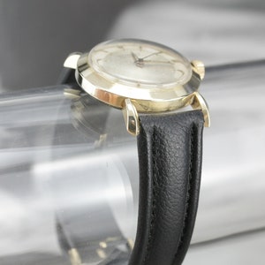 Pu&ograve; includere: Un orologio da polso dorato con quadrante bianco e cinturino in pelle nera. L'orologio ha una cassa rotonda e un design semplice.