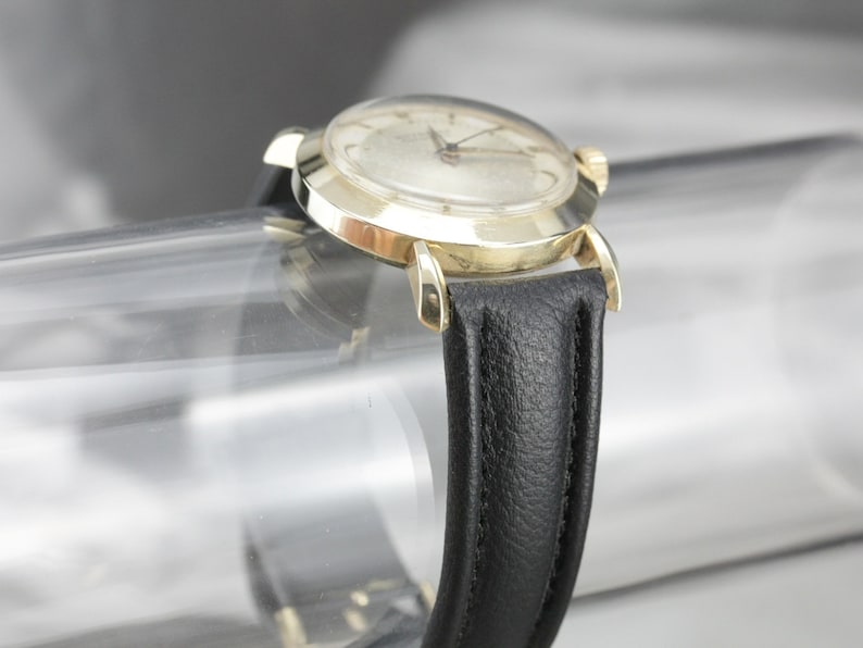 Pu&ograve; includere: Un orologio da polso dorato con quadrante bianco e cinturino in pelle nera. L'orologio ha una cassa rotonda e un design semplice.