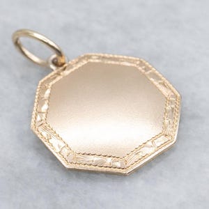 May include: A gold-coloured, octagonal pendant with a decorative border and a small loop at the top. The pendant has a smooth, blank surface, suggesting it could be engraved or personalised.