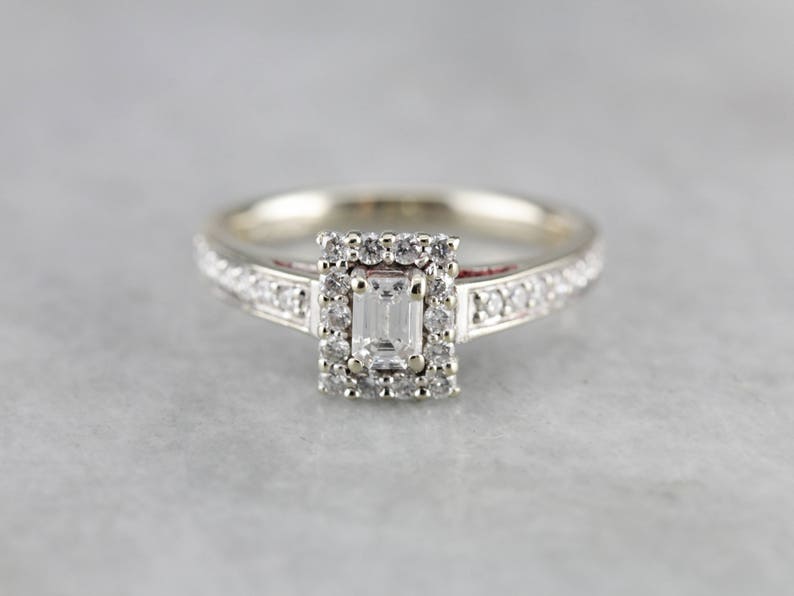 Puede incluir: Un anillo de plata con un diamante talla esmeralda de forma cuadrada engastado en un halo de diamantes m&aacute;s peque&ntilde;os. La banda tambi&eacute;n est&aacute; engastada con diamantes m&aacute;s peque&ntilde;os.