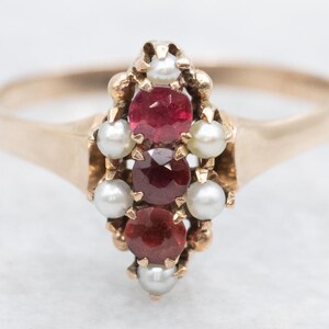 May include: A gold ring featuring a cluster of three red gemstones and six small white pearls. The ring's band is a simple, polished gold, and the gemstones are set in a decorative gold setting.