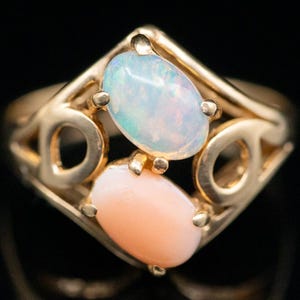 Puede incluir: Un anillo de oro con dos opales de forma ovalada. El ópalo más grande es blanco con un toque de rosa, y el ópalo más pequeño es rosa pálido. El anillo tiene un diseño delicado e intrincado.