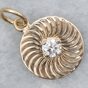 May include: A circular gold pendant featuring a central round brilliant-cut diamond set in a four-prong mount. The surface of the pendant is decorated with a radiating spiral pattern of curved grooves that extend from the center to the edge.