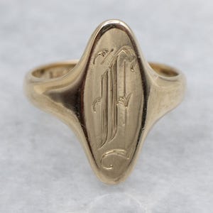 Puede incluir: Un anillo de sello dorado con una cara ovalada. La cara del anillo presenta un monograma grabado en una fuente estilizada. El anillo es de color oro cálido y tiene un acabado liso y pulido.