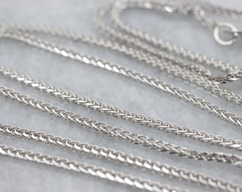 May include: Close-up of a silver wheat chain necklace. The chain is made of small, interwoven links, creating a textured surface. The necklace is displayed on a light gray surface, with the chain winding across the frame. Jewelry.