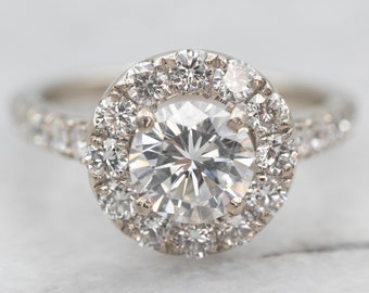 Könnte beinhalten: Ein silberner Ring mit einem großen runden Diamanten in der Mitte, umgeben von einem Halo aus kleineren Diamanten. Die Ringschiene ist ebenfalls mit kleineren Diamanten besetzt.