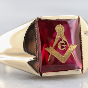 May include: A gold ring with a large, square, red gemstone. The gemstone has a gold Masonic symbol engraved on it.
