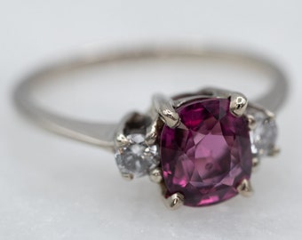 May include: A silver ring with a large, dark pink gemstone surrounded by two smaller, clear gemstones.