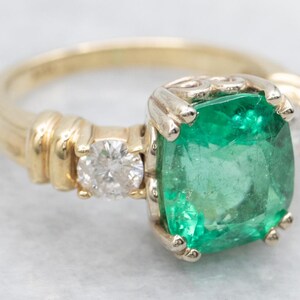 May include: A gold ring featuring a large, square-cut emerald gemstone. A smaller diamond is set on either side of the emerald. The ring has a detailed band with a ribbed design.