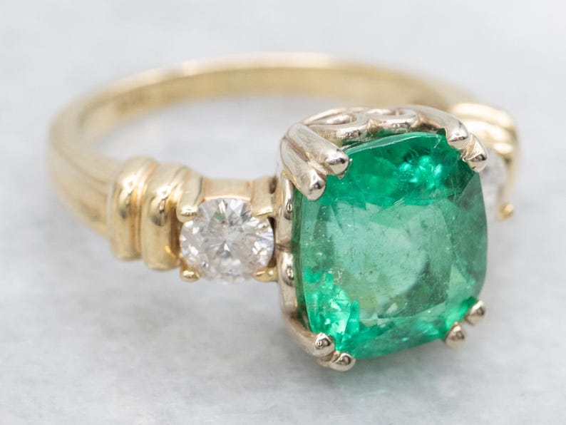 May include: A gold ring featuring a large, square-cut emerald gemstone. A smaller diamond is set on either side of the emerald. The ring has a detailed band with a ribbed design.