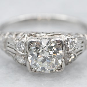Puede incluir: Primer plano de un anillo de oro blanco con un gran diamante central de talla cuadrada. El anillo presenta intrincados detalles de filigrana y pequeños diamantes de acento en la banda. El fondo es neutro.