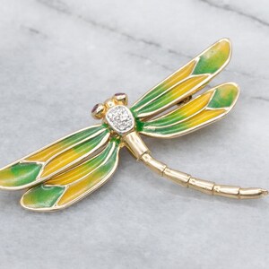 May include: A gold-toned dragonfly brooch with green and yellow enamel wings. The dragonfly's head features a cluster of diamonds and two small red gemstones. The brooch is a detailed, elegant piece of jewelry.
