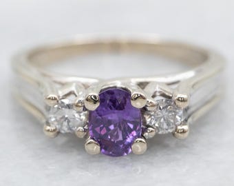 May include: A white gold ring with a central oval-cut purple gemstone, flanked by two smaller, round, clear gemstones. The ring has a classic three-stone design, with a polished band and prong settings.
