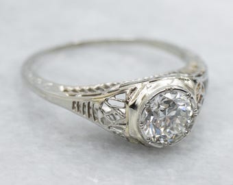 May include: A vintage-style ring with a large, round, clear diamond set in a white metal band. The band features intricate filigree detailing on the sides and a smooth, polished finish. The ring is set against a soft, neutral background.