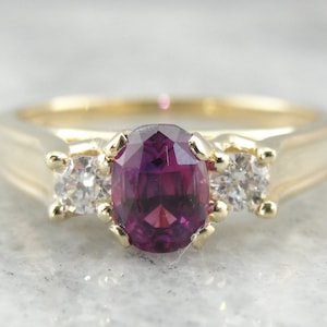 May include: A gold ring featuring a large oval purple gemstone flanked by two smaller round white gemstones.