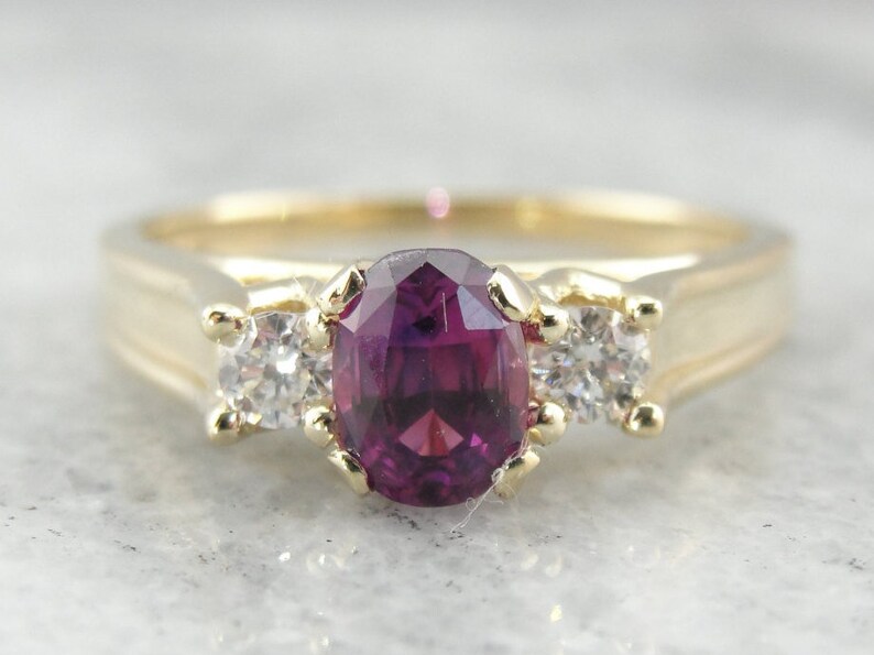 May include: A gold ring with a large oval-shaped purple gemstone surrounded by two smaller round white gemstones.