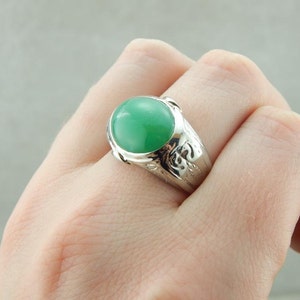 May include: A silver ring with a green gemstone set in a bezel setting. The ring has a detailed design around the gemstone.