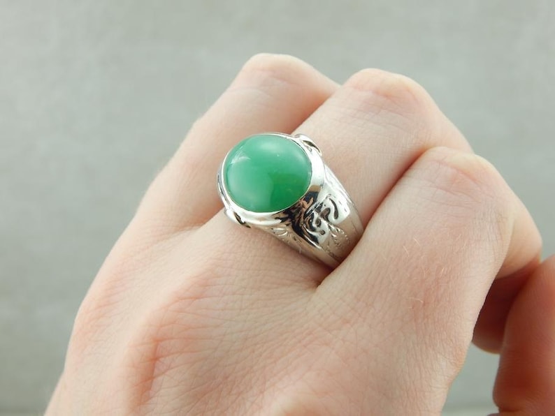 May include: A silver ring with a green gemstone set in a bezel setting. The ring has a detailed design around the gemstone.