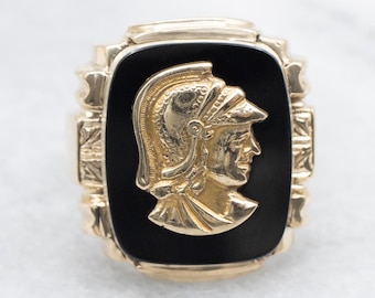 May include: A gold ring set with a black onyx stone, featuring a detailed, gold-toned profile of a person wearing a helmet.