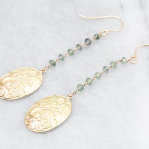 Puede incluir: Pendientes de oro con una cadena delicada de piedras preciosas verdes y colgantes ovalados con un diseño floral intrincado.