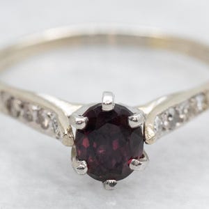 Puede incluir: Un delicado anillo de oro con una piedra preciosa roja oscura rodeada de pequeños diamantes. El anillo tiene una banda simple con un diseño delicado.