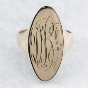 Op de afbeelding: Goudkleurige ovale zegelring met een gegraveerd monogram. De ring heeft een gepolijste afwerking en een klassiek ontwerp. Het monogram is ingewikkeld gedetailleerd, wat een persoonlijk tintje aan het sieraad geeft.