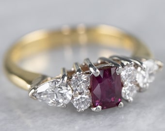 May include: A gold and silver ring with a large red ruby gemstone surrounded by smaller diamonds. The ring has a pear-shaped diamond on either side of the ruby.