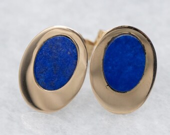 Può includere: Un paio di gemelli ovali color oro con un'intarsio di pietra blu vibrante. Il metallo lucido incornicia la pietra blu profonda e strutturata, creando un accessorio classico ed elegante. I gemelli sono presentati su una superficie chiara.