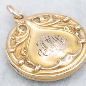 May include: A round gold locket pendant featuring an ornate, raised scrollwork border surrounding a smooth, polished center. The center is engraved with an intricate, cursive monogram. A small gold jump ring is attached to the top.