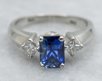 May include: A silver ring featuring a large, rectangular, deep blue gemstone as the centerpiece. Two smaller, square, clear gemstones flank the central stone. The ring has a classic, elegant design.