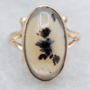 May include: A gold ring featuring an oval-shaped, translucent stone with black dendritic inclusions. The ring has a split shank design and a decorative setting. The stone's dimensions are approximately 1 inch in length and 0.75 inches in width.